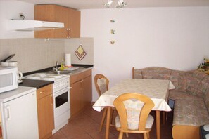 Een kleine keuken met een fornuis, magnetron en spoelbak, een eettafel met stoelen, en een bank in de hoek.
