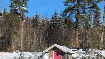 A small wooden cabin with a pink door, surrounded by snow-covered ground and tall pine trees.