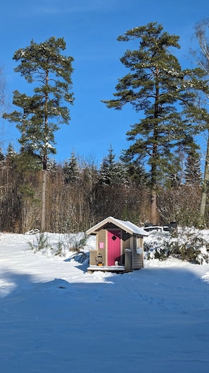 A small wooden cabin with a pink door, surrounded by snow-covered ground and tall pine trees.
