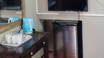 A hotel room with a bed, a small table with a tray of cups, a television mounted on the wall, and a compact refrigerator.