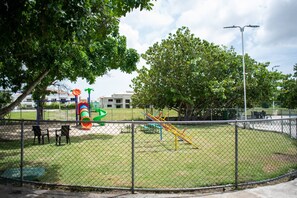 Ein eingezäunter Spielplatz mit Spielgeräten, Bänken und einem Gebäude im Hintergrund.