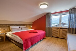 Une chambre avec une tête de lit en bois, un lit recouvert d’une couverture rouge, une table de chevet, une fenêtre avec des rideaux et une vue sur les montagnes.