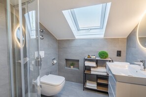 Une salle de bain moderne dotée d’un puits de lumière, d’une douche à parois de verre, de toilettes, d’un lavabo et d’une étagère où sont rangées des serviettes.