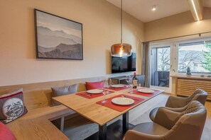 Un coin repas avec une table en bois dressée pour quatre personnes, un canapé avec des coussins, un téléviseur fixé au mur et une fenêtre donnant sur la forêt.