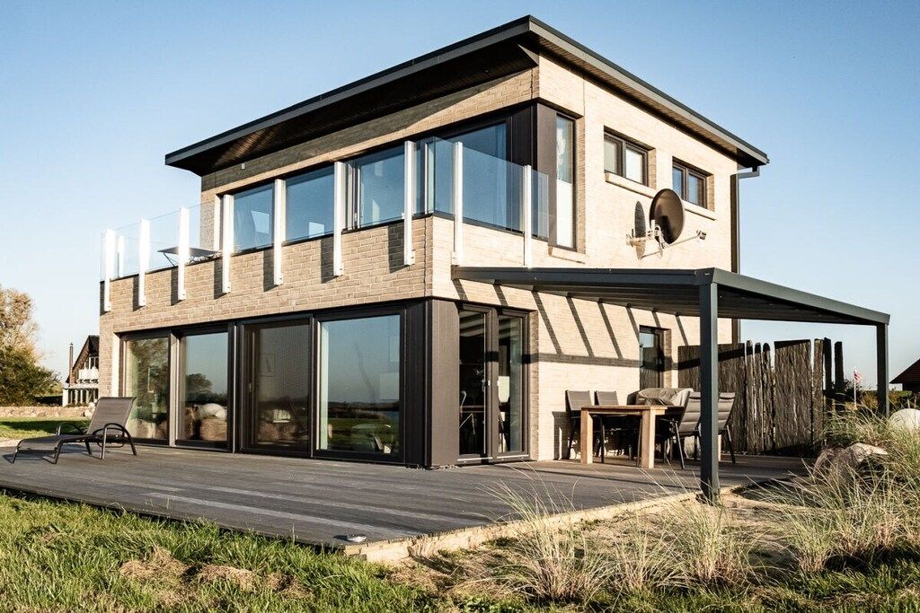 A modern two-story house with large glass windows, a wooden deck, and outdoor furniture.