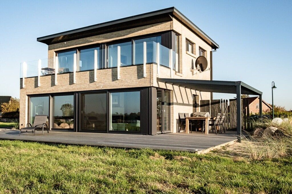 A modern two-story house with large glass windows, a wooden deck, and a covered outdoor seating area.