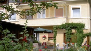 A two-story building with a covered outdoor seating area, a garden with various plants and flowers, and a bicycle parked outside. - Holiday apartment with 1 bedroom (Traben-Trarbach)