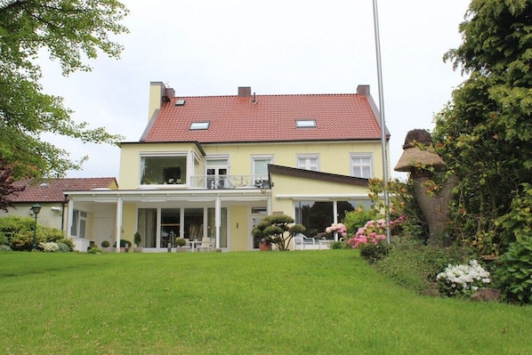 Une maison à deux étages dotée d’un porche abrité et d’un jardin où poussent diverses plantes et fleurs.