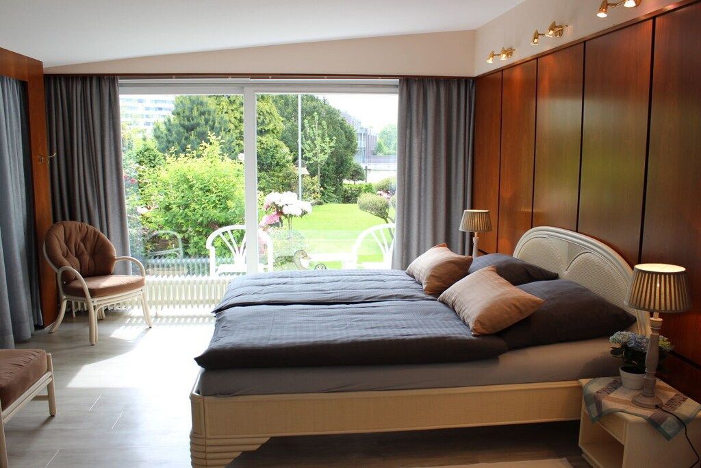 A bedroom with a bed, a chair, a lamp, and a view of a garden through a large window.