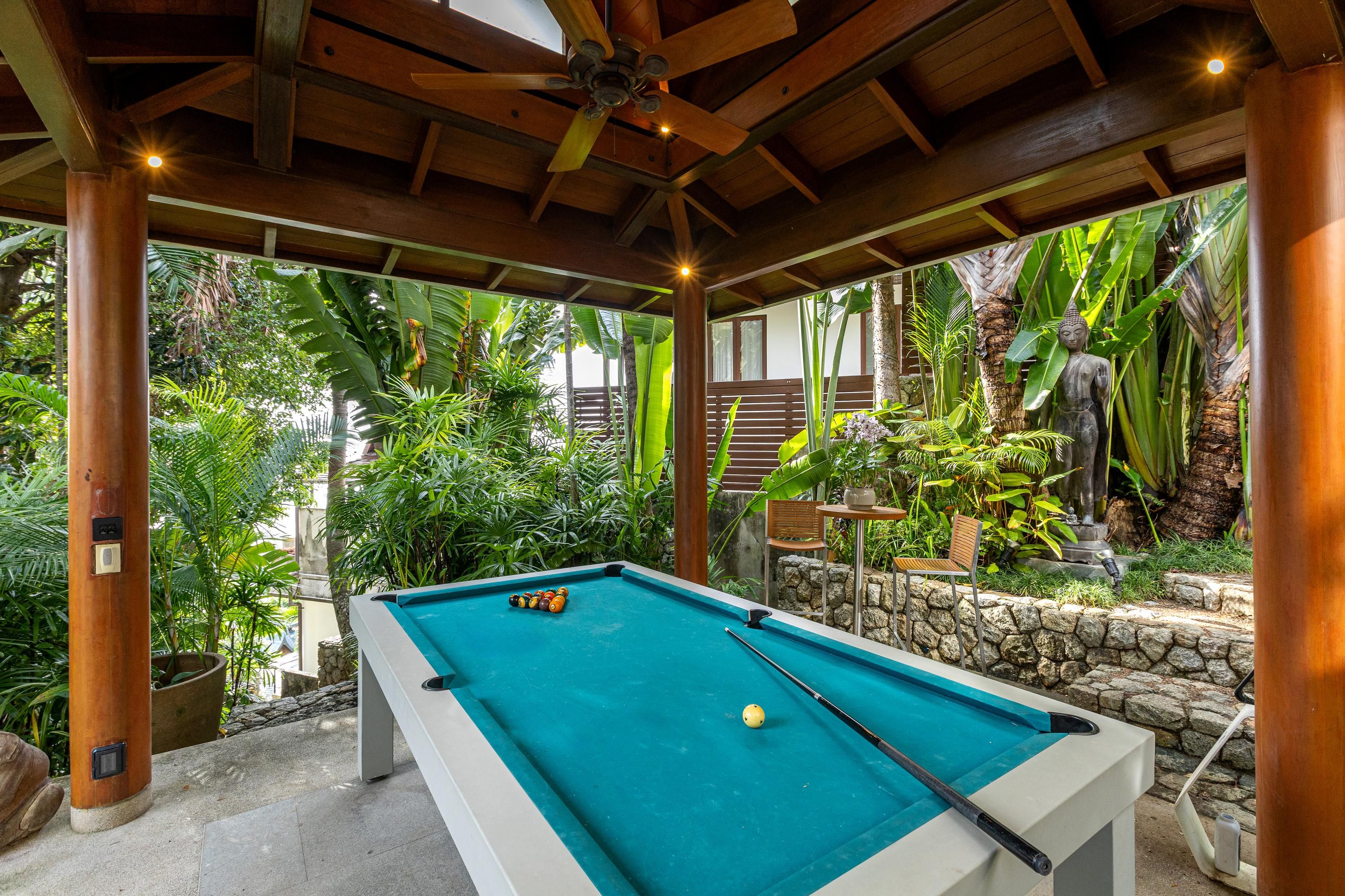 Un espace piscine intérieur doté d’une table de billard, entouré d’une végétation tropicale luxuriante et surmonté d’une structure en bois.