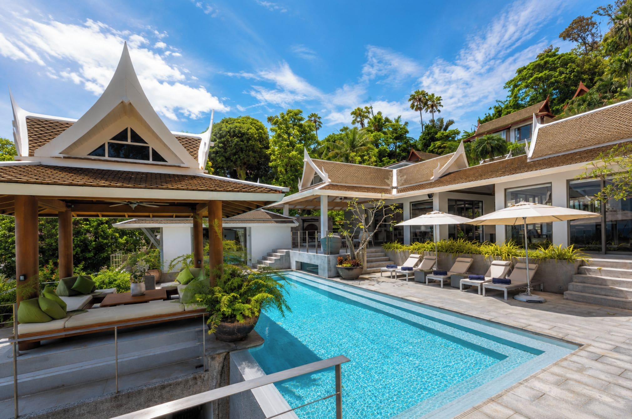 A swimming pool with lounge chairs and a covered seating area, surrounded by lush greenery and traditional architecture.