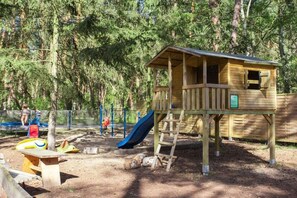 Eine hölzerne Spielanlage mit Rutsche und einem kleinen hausähnlichen Gebäude in einem bewaldeten Park.