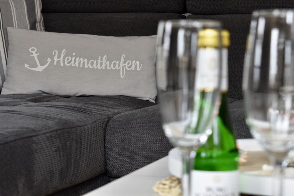 Ein graues Sofa mit einem Kissen, auf dem 'Heimathafen' steht. Vor dem Sofa befinden sich zwei Weingläser und eine Weinflasche.