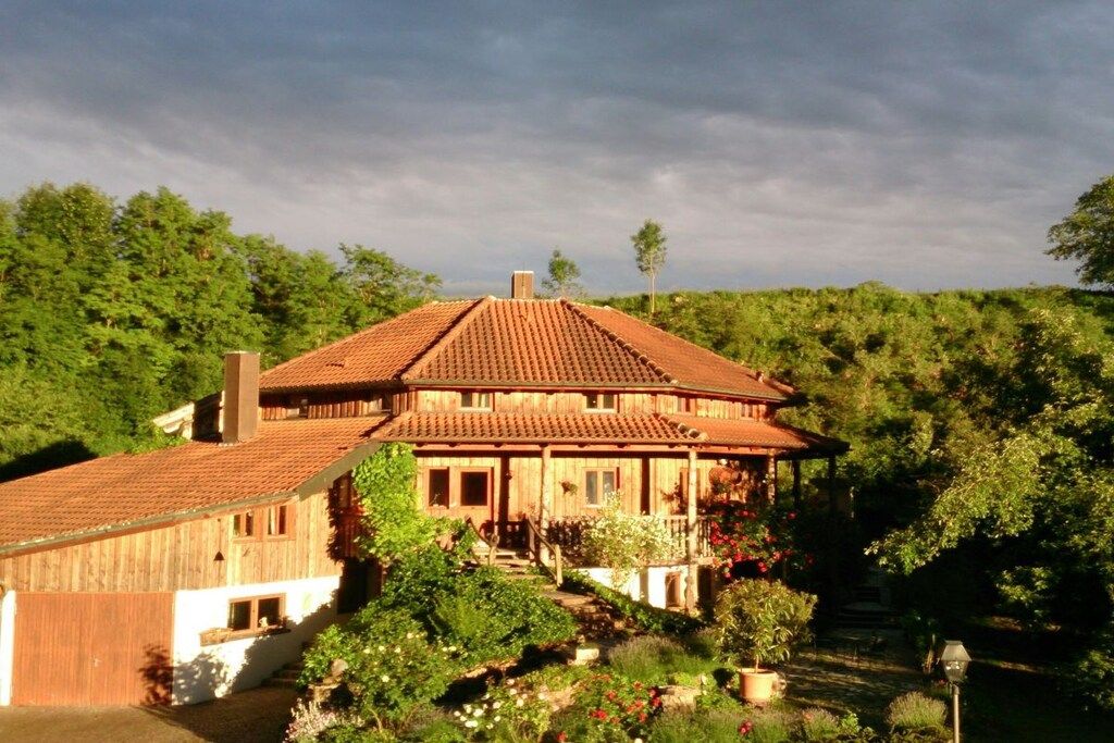 A two-story wooden house with a tiled roof, surrounded by lush greenery and trees.