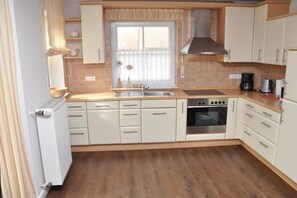 A kitchen with white cabinets, a stove, a sink, and a window with blinds. - Holiday apartment with 1 bedroom (Krummhörn \/OT Greetsiel)