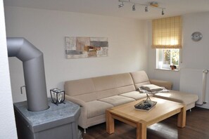 A living room with a sofa, a coffee table, a fireplace, a window with blinds, and a clock on the wall. - Holiday apartment with 1 bedroom (Krummhörn \/OT Greetsiel)