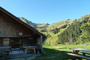 A rustic mountain lodge with a wooden exterior, a small porch, and picnic tables in a valley surrounded by mountains. - Iseler - Landhaus Thomma (Oberjoch)