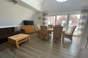 Ein Wohnzimmer mit einer Couch, einem Couchtisch und Korbstühlen um einen Tisch herum. Ein Fernseher steht auf einem Schrank, und es gibt ein Fenster mit Vorhängen.