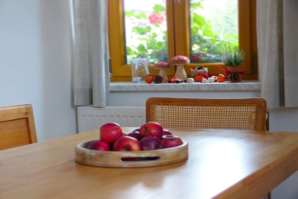 Et træbord med en skål æbler, en fletstol og et vindue med planter og udsigt til grøn natur.