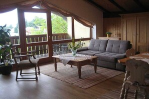 A cozy living room with a sofa, a wooden coffee table, and a chair. There is a balcony with a railing and a view of greenery outside. - Holiday apartment with 1 bedroom (Wegscheid)