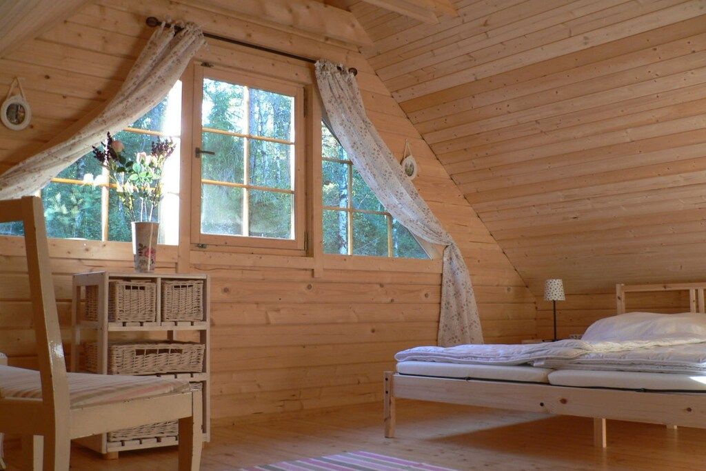 A wooden cabin room with a bed, a chair, a small table, and a window with curtains.