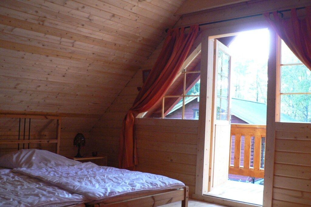 A wooden cabin bedroom with a bed, a nightstand, a window with curtains, and a view of the outside.