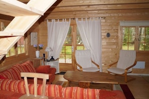 A cozy cabin interior with a wooden ceiling, a sofa, two chairs, a small table, and a window with curtains.