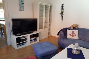 A living room with a flat-screen TV on a stand, a glass cabinet, a blue sofa with an anchor pillow, and a small table with a decorative item. - Holiday home with 1 bedroom (Visselhövede)
