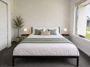 A neatly arranged bedroom with a bed, two bedside tables, a potted plant, and a window. - The Olive Nest - Modern Family Comfort (Upper Coomera)
