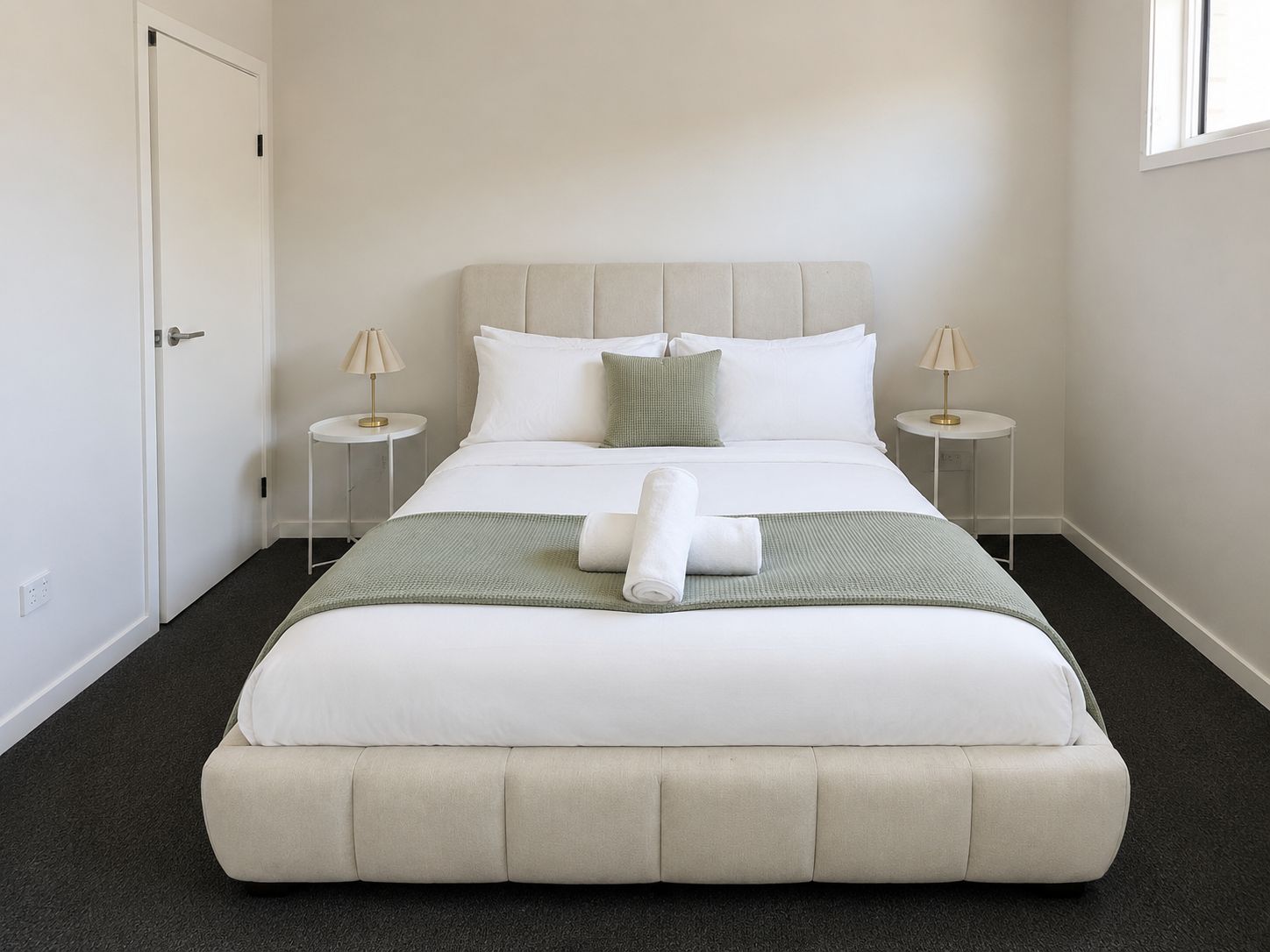 A neatly made bed with white bedding and a green throw, two bedside lamps, and a door to the left.