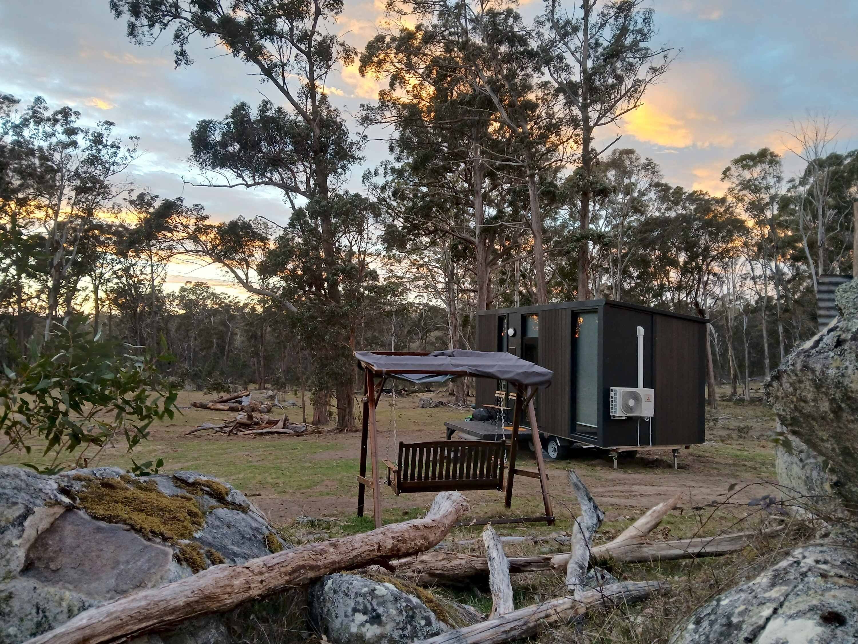 A small, dark-colored cabin with a bench and a table, surrounded by trees and rocks.