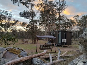 A small, dark-colored cabin with a bench and a table, surrounded by trees and rocks.
