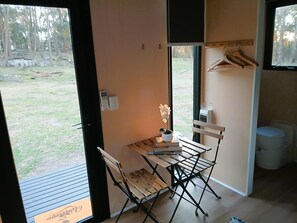 A room with a wooden table, chairs, a bench, a hanging clothes rack, and a window with a view of a grassy area.