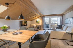 A modern living room with a wooden ceiling, a sofa, a dining table with chairs, and a large window with curtains. - Holiday apartment in Oberstdorf with balcony (Oberstdorf)