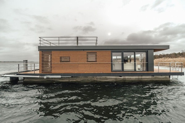 A modern floating house with a wooden deck, large windows, and a metal railing.
