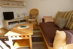 A living room with a television, a wooden table, a wicker chair, a sofa, and a striped cushion. - Holiday apartment with 1 bedroom (Bernau am Chiemsee)