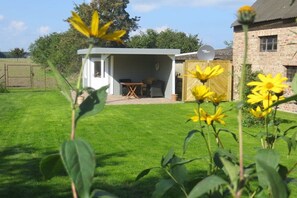 Ein kleines, eingeschossiges Gebäude mit einer überdachten Sitzterrasse, ein Backsteinhaus im Hintergrund und ein gepflegter Rasen mit gelben Blumen.