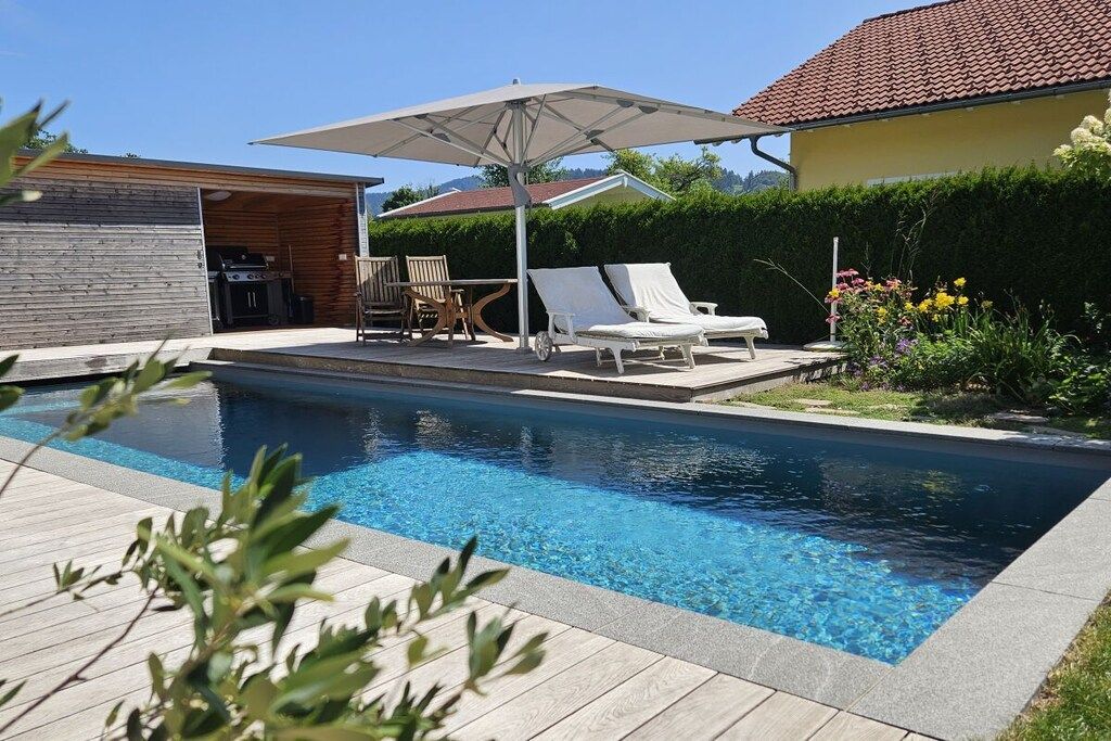 A swimming pool with lounge chairs and an umbrella, surrounded by a wooden deck and a garden.