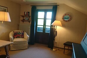 Ein Zimmer mit einem gemusterten Sessel, einem kleinen Tisch, einer Stehlampe, einem Heizkörper und einem Fenster mit Vorhängen.