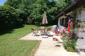 A patio with a table and chairs, a covered umbrella, and a white building with a garden. - Holiday home on Lake Chiemsee (Breitbrunn am Chiemsee)