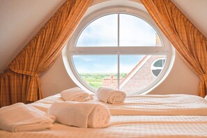 A bedroom with a large round window, a bed with white linens, and a view of a brick building. - Holiday apartment with 1 bedroom (Krummhörn \/OT Greetsiel)