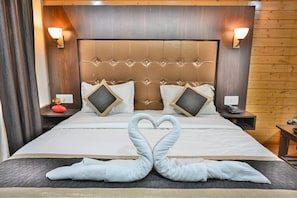 Quarto de hotel com uma cama grande, duas mesas de cabeceira, um cabeceira de madeira e uma decoração de toalha em formato de coração.