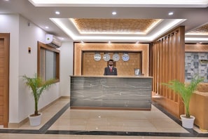 Recepção de hotel com balcão de mármore, relógios e plantas em vasos.