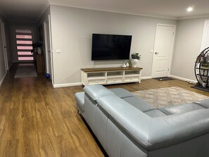 A modern living room with a grey leather sofa, a wooden entertainment unit, and a flat-screen TV mounted on the wall.