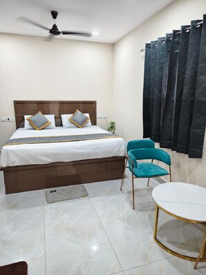 Un dormitorio con cama, ventilador de techo, ventana con cortinas, una silla y una mesita.