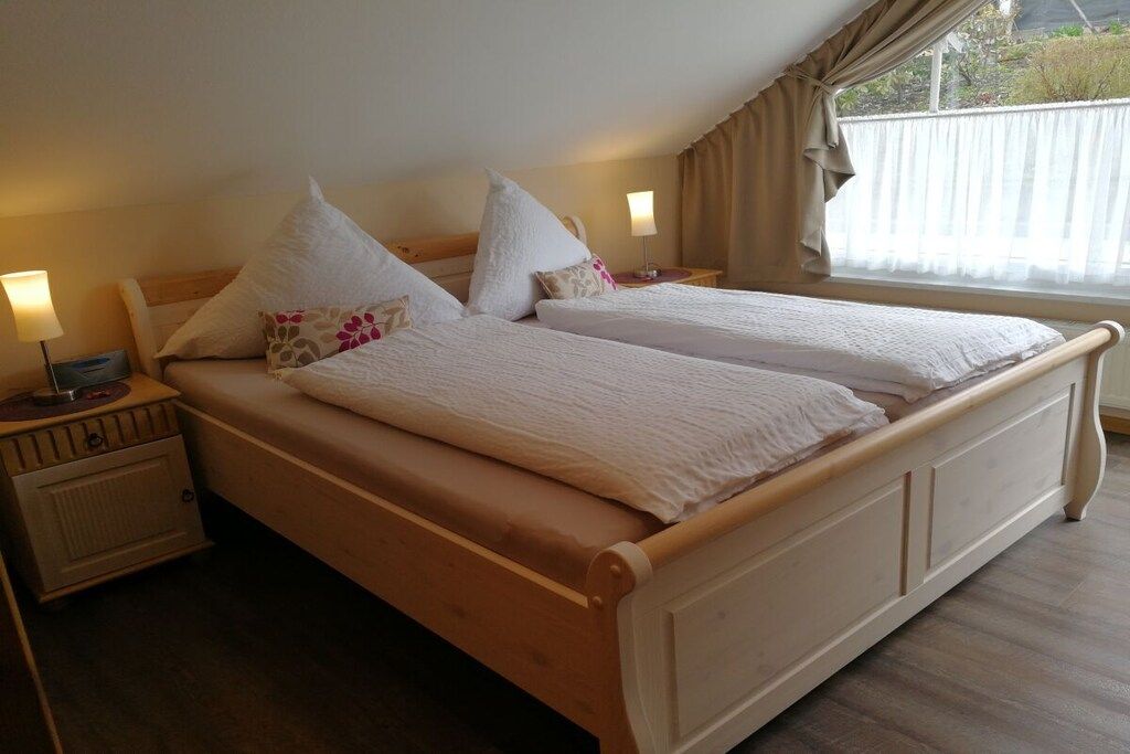 Ein Doppelbett mit Holzrahmen und weißer Bettwäsche, ein Nachttisch mit Lampe sowie ein Fenster mit Vorhängen.