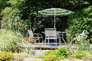 An outdoor seating area with a table and chairs, a green and white umbrella, and a garden with various plants. - Holiday apartment with 1 bedroom (St. Märgen)