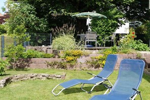 Zwei Liegestühle, ein Tisch mit Sonnenschirm und ein Garten mit unterschiedlichen Pflanzen und einem Zaun.