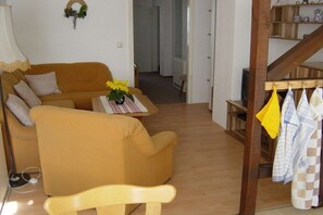 Un salon comprenant un canapé, une table basse ornée de fleurs, une chaise en bois et un escalier sur lequel sont suspendus des torchons de cuisine.