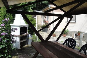 Un patio couvert avec une table et des chaises en bois, un four d’extérieur blanc et des plantes en pot.